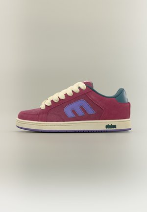 Niedriger Sneaker aus rosa Wildleder mit cremefarbenen Schnürsenkeln, lila Logo auf der Seite, grünem Absatz und cremefarbener Sohle mit lila Detail auf neutralem Hintergrund.