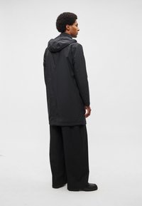 Fekete vízálló kapucnis dzseki középhosszú szabással, sima textúrával és minimalista stílussal, széles szárú fekete nadrággal párosítva.