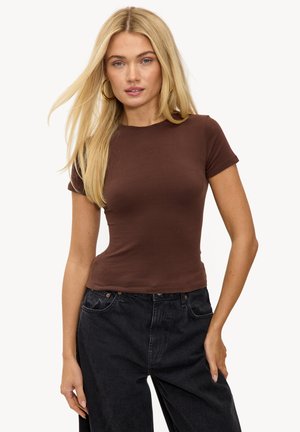 Blonde vrouw met lang haar, draagt een nauwsluitend bruin kort mouwtopje en een zwarte spijkerbroek met hoge taille, staand tegen een witte achtergrond.