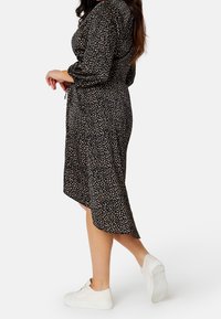 Schwarzes Kleid mit einem verstreuten beigen Punktmuster, langen Ärmeln, einer taillierten Taille und einem asymmetrischen Saum, kombiniert mit weißen Sneakern.
