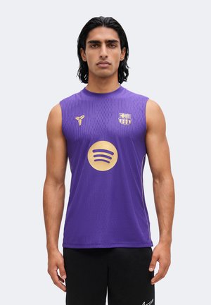 Homme aux cheveux noirs mi-longs portant un maillot de sport violet sans manches avec les logos du FC Barcelone et de Spotify, debout devant un fond uni.