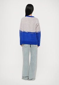 Asmuo atsisukęs atgal, vilkintis laisvą mėlyną ir pilką gradientinį megztinį bei plačias šviesias džinsines kelnes prieš paprastą foną.