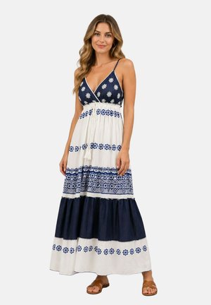 Femme portant une robe maxi sans manches bleu marine et blanche à rayures horizontales à motifs et des sandales marron, debout et souriante.