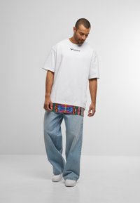 Bărbat îmbrăcat cu un tricou alb Colucci peste o cămașă lungă cu un imprimeu colorat, blugi deschiși la culoare, lejeri, și adidași albi, stând pe un fundal simplu.