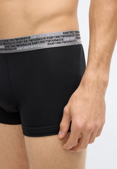 Fekete, rugalmas anyagból készült boxeralsó szürke derékrésszel, amelyen az "EMPORIO ARMANI" márkajelzés látható, sima felülettel. Oldalnézetből mutatva.