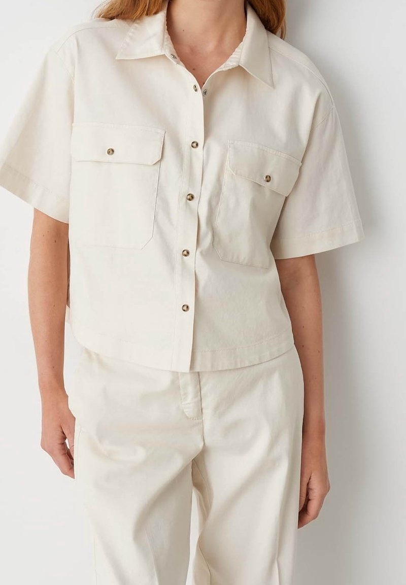 Chemise à manches courtes de couleur crème avec col et deux poches poitrine. Fabriquée en coton, dotée de boutons en métal et d'une coupe décontractée.