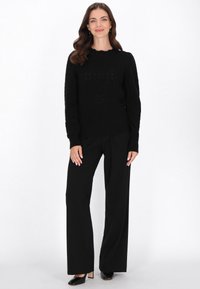 Czarny sweter dzianinowy o teksturowanym wzorze, z falbaniastym dekoltem; zestawiony z czarnymi spodniami o szerokich nogawkach i czarnymi butami na obcasie.