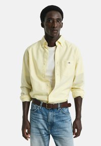 Geel gestreepte blouse met een borstzak, gelaagd over een wit T-shirt, in combinatie met blauwe spijkerbroeken en een bruine riem.