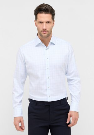 Man die een lichtblauw overhemd met lange mouwen en donkere broek draagt, staand tegen een effen witte achtergrond.