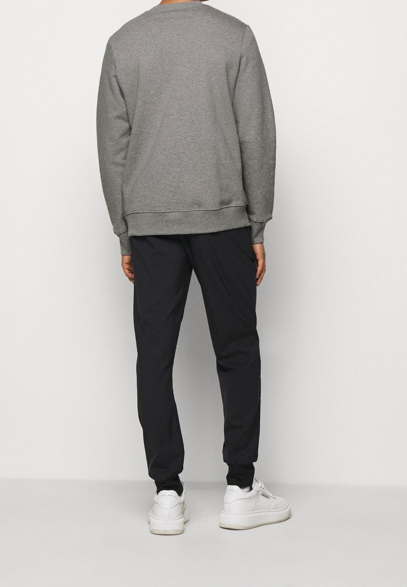 Personne portant un sweat-shirt gris à manches longues, un pantalon de jogging noir et des baskets blanches, debout de dos devant un fond uni.