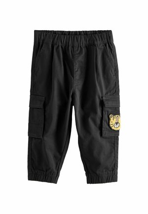 Enfant portant un t-shirt à manches courtes moutarde, un pantalon cargo noir avec un écusson tigre, et des baskets blanches à velcro, debout devant un fond beige.