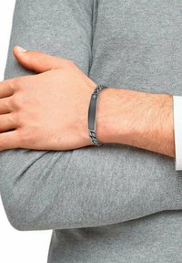 Gray braided bracelet with a textured black metal accent. The metal section features a grid pattern and a central insignia.