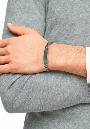 Grau geflochtener Armband mit einem strukturierten schwarzen Metallelement. Der Metallbereich zeigt ein Rautenmuster und ein zentrales Symbol.