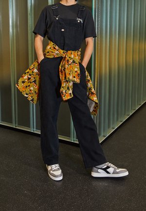 Osoba ubrana w czarne ogrodniczki, czarną koszulkę, szaro-białe trampki, z kurtką w kamuflażu zawiązaną wokół talii, stojąca wewnątrz pomieszczenia.