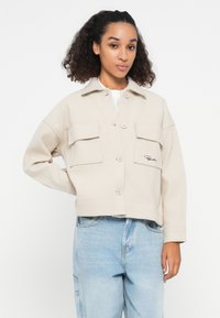 Cropped beige jacket with a collar, four front pockets, and button closure. Smooth texture. Paired with light blue jeans.