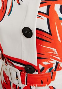 Tissu blanc avec des motifs abstraits audacieux en rouge, orange et noir ; présente des boutons noirs et une ceinture, soulignant le design ajusté.