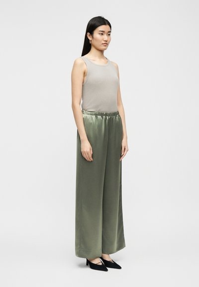 Γυναίκα που στέκεται με μπεζ αμάνικο τοπ, σατέν, ελιάς πράσινο παντελόνι φαρδιάς γραμμής και μαύρα μυτερά τακούνια, μπροστά σε απλό ανοιχτό φόντο.
