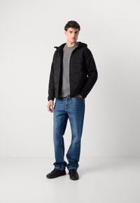 Zwarte gewatteerde jas met capuchon, grijze ribbeltrui, blauwe rechte spijkerbroek en zwarte sneakers. Model staat op een witte achtergrond.