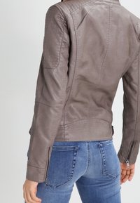 Chaqueta de cuero gris con acentos texturizados en los hombros, puños con cremallera y diseño ajustado. Llevada sobre jeans azules, mostrando la vista trasera.