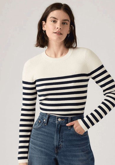 Woman wearing a white sweater with black horizontal stripes and high-waisted blue jeans, posing with one hand in her pocket.