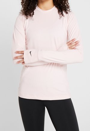 Femme aux cheveux bouclés portant un haut Nike rose clair à manches longues avec des trous pour les pouces et des leggings noirs, bras croisés sur la poitrine.