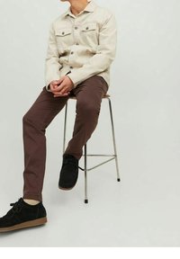 Giubbotto di jeans beige con due tasche sul petto, pantaloni marroni a sigaretta e scarpe in suede nere su uno sgabello di metallo contro uno sfondo chiaro.