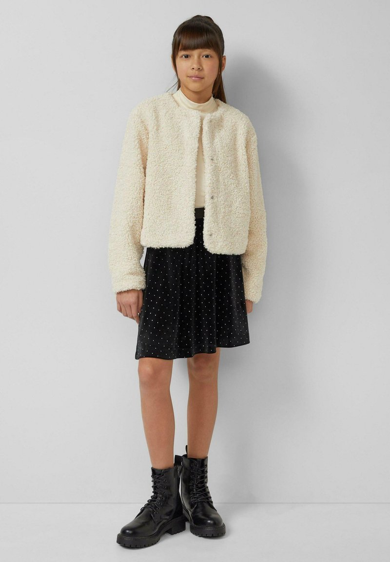 Kremowa kurtka z futerkiem ekologiczny, z okrągłym dekoltem, zestawiona z czarną aksamitną spódnicą w białe kropki. Modelka ma na sobie czarne sznurowane botki.