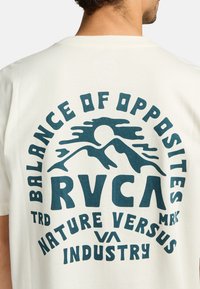 Cremefarvet bomulds t-shirt med et blåt grafisk design, der viser bjerge, en sol og teksten "BALANCE OF OPPOSITES RVCA NATURE VERSUS INDUSTRY."