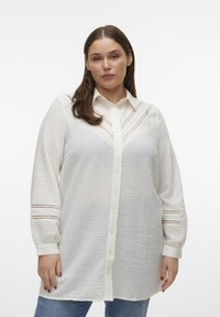 Camisa blanca de botón en tela texturada ligera, con intrincados detalles recortados en los hombros y las mangas, con un corte relajado.