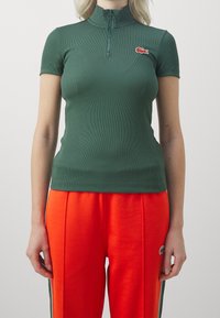 Zelené žebrované polo tričko s krátkým rukávem a zipovým krkem, s logem Lacoste na hrudi, v kombinaci s oranžovými sportovními kalhotami s bočními pruhy.