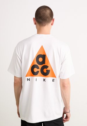 Vīrietis ar īsiem matiem, tērpies baltajā T-kreklā ar lielu oranžu trifeli un melnajiem "acg" burtiem virs "NIKE" uz muguras.
