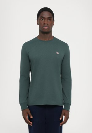 Mann, der ein dunkelgrünes Langarmshirt mit einem kleinen bunten Elefantenlogo auf der linken Brust trägt, vor einem schlichten weißen Hintergrund steht.