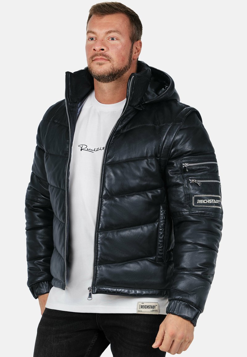 Giubbotto imbottito di colore blu scuro, con cappuccio, chiusura con zip, pannelli cuciti e toppe con logo sulle maniche. Indossato sopra una maglietta bianca.