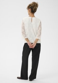 Hvid blondebluse med lange, gennemsigtige ærmer og nøglehulslukning i ryggen. Parret med løstsiddende, sorte bukser. Glat, struktureret stof.