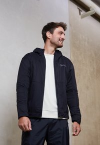 Giacca leggera nera con cappuccio, dotata di una chiusura lampo frontale, pannelli testurizzati e un logo sul petto. Indossata sopra un maglione color crema.