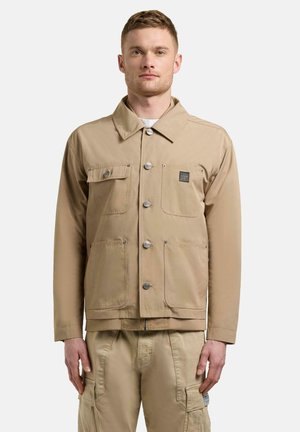 Mężczyzna ubrany w beżową kurtkę zapinaną na guziki z czterema kieszeniami z przodu oraz dopasowane beżowe spodnie typu cargo, stojący na jednolitym tle.