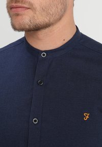 Man in een donkerblauw overhemd met mandarin collar, voorzien van drie zwarte knopen en een klein oranje geborduurd 'F'-logo op de borst.