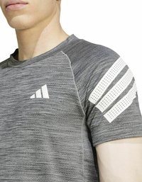 Camiseta atlética gris con un patrón texturizado y cuello redondo. Presenta acentos blancos, incluyendo el logo y un diseño a rayas en el hombro.