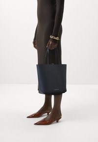 Marinblå läderaxelväska med två handtag, minimalistisk design. Har en liten logotyp fram, tillsammans med bruna spetsiga klackskor.