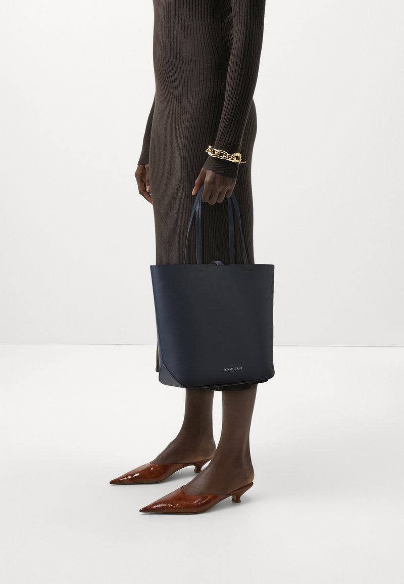 Marinblå läderaxelväska med två handtag, minimalistisk design. Har en liten logotyp fram, tillsammans med bruna spetsiga klackskor.