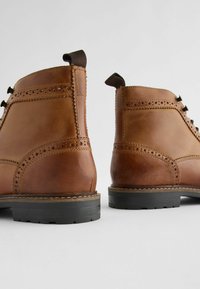 Botas de cuero marrón hasta el tobillo con detalles de brogue, cordones planos y suelas de goma negra. Textura suave con acentos de costura en contraste.