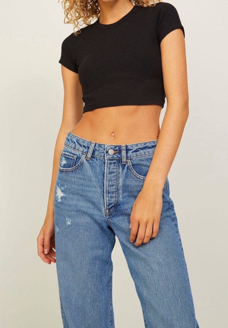 Woman wearing a black cropped t-shirt and high-waisted blue jeans with slight distressing, standing against a plain light background.