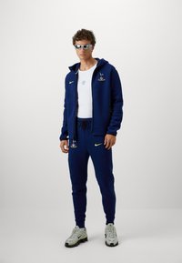 Mørkeblå træningsdragt med broderede logoer, hættejakke med fuld lynlås og elastisk talje; parret med grå sneakers med sorte detaljer.