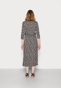 Vrouw die met haar rug naar de camera staat, gekleed in een zwarte midi-jurk met bloemenprint, driekwartmouwen en zwarte sandalen.