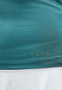 Κοντινό πλάνο σε πετρόλ ύφασμα με ραμμένο τελείωμα και ανάγλυφο κείμενο "BORG" πάνω από λευκό ύφασμα από κάτω.