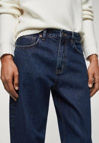 Jeans di denim blu scuro a gamba dritta, con tasche frontali e dettagli di cucitura sottili, abbinati a un maglione lavorato a maglia di colore crema.