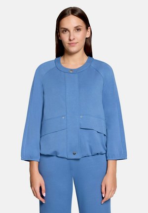 Frau trägt eine blaue langärmlige Jacke mit Druckknöpfen und passende Hose, steht vor einem schlichten Hintergrund.