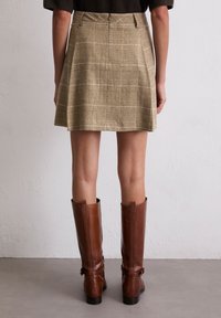 Mini-jupe à carreaux marron avec des plis, fabriquée en tissu texturé. Portée avec des bottes en cuir marron jusqu'au genou avec un détail de sangle.