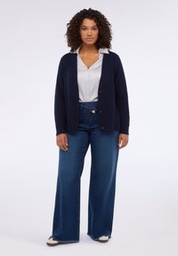 Námořnický pletený kardigan s knoflíky, který je navrstvený přes světle šedou košili. Model má na sobě široké tmavě modré džíny a casual tenisky.