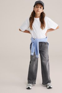 Bela prevelika majica, sive široke kavbojke, modri pulover, zavezan okoli pasu, siva kapa in superge z teksturirano podplatom.